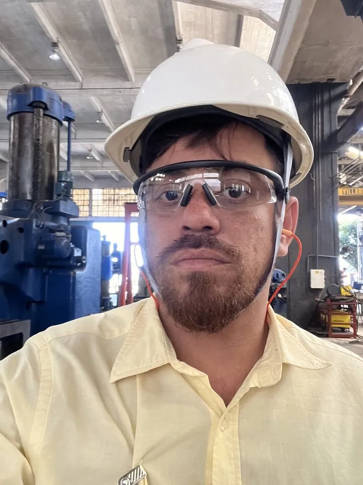 Técnico com capacete branco e óculos de proteção dentro de uma fábrica, com máquinas ao fundo